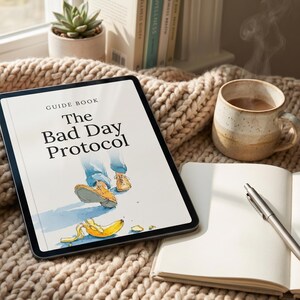 Puede incluir: Una tableta muestra la guía "The Bad Day Protocol", con una ilustración de pies y una cáscara de plátano. Una taza de café, un cuaderno y un bolígrafo están sobre una manta de punto. Una pequeña planta suculenta es visible.
