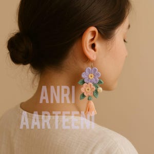 Könnte beinhalten: Handgefertigte Häkelohrringe mit floralem Design, darunter eine lila Blume, eine pfirsichfarbene Blume und grüne Blätter. Die Ohrringe haben eine pfirsichfarbene Quaste und goldfarbene Haken. Der Text "ANRI AARTEEN" ist sichtbar.