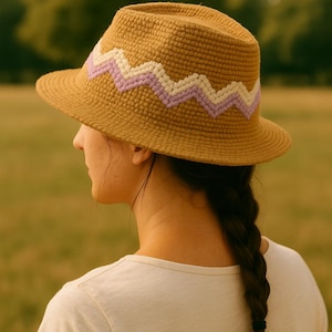 May include: A woven fedora-style hat in a tan color, featuring a zigzag pattern in white and lavender around the crown. The hat has a wide brim and is worn by a person with a braid.