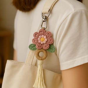 Puede incluir: Un llavero de flores de ganchillo hecho a mano con una flor rosa y amarilla, hojas verdes y una borla beige. El llavero está sujeto a un bolso tote beige con una correa color crema. La flor mide aproximadamente 7.6 cm de diámetro.