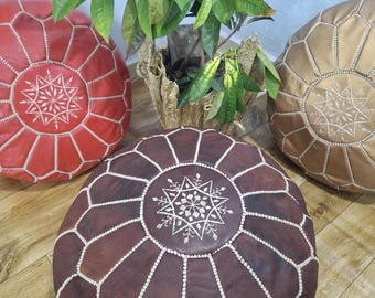 Può includere: Tre pouf marocchini in pelle rossa, marrone e oro. Ogni pouf rotondo presenta un design a stella geometrica con cuciture bianche al centro e lungo le cuciture. Una pianta da appartamento è sullo sfondo.