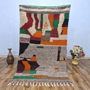 May include: A large, rectangular rug with a colourful, abstract design. The rug features a mix of colours, including grey, orange, red, green, and white. The rug has a fringe along the bottom edge and is displayed indoors.