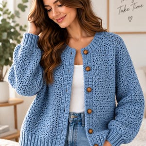 May include: A light blue crochet cardigan featuring a round neckline, long sleeves with ribbed cuffs, and a front button closure with six brown wooden buttons. The garment is worn over a white top and light blue denim jeans.