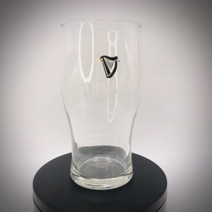 May include: A clear pint glass with the Guinness logo, a black harp with a gold accent, is displayed. The glass has a curved shape and a wide mouth. The Guinness logo is printed on the front of the glass.
