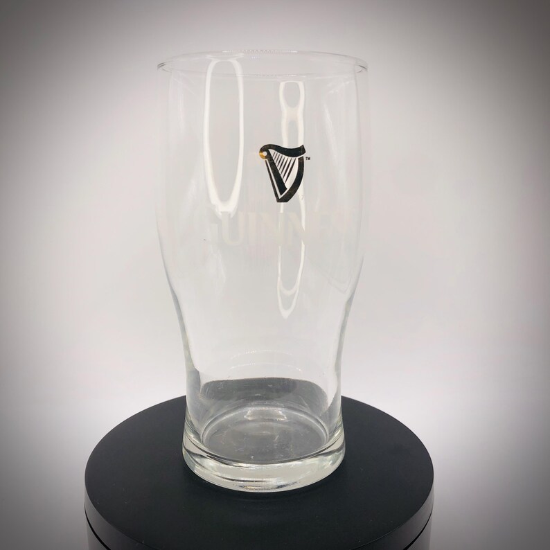 May include: A clear pint glass with the Guinness logo, a black harp with a gold accent, is displayed. The glass has a curved shape and a wide mouth. The Guinness logo is printed on the front of the glass.