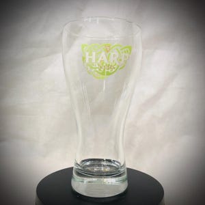 May include: A clear glass beer pilsner with the Harp Lager logo in green and white. The logo features the word "HARP" above "LAGER" with a Celtic knot design. The glass is on a black surface.