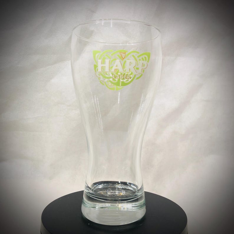 May include: A clear glass beer pilsner with the Harp Lager logo in green and white. The logo features the word "HARP" above "LAGER" with a Celtic knot design. The glass is on a black surface.