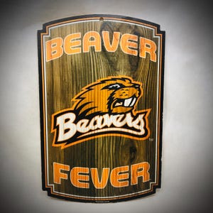 May include: Wooden sign with a wood grain background featuring the words "BEAVER FEVER" in orange lettering. The sign also displays a cartoon beaver mascot in orange and black. The sign has a black border and a hole for hanging.