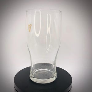 May include: A clear, pint-sized glass with a slightly curved shape. The glass features a gold harp logo and the word "Guinness" printed vertically. The glass is empty and sits on a black surface.
