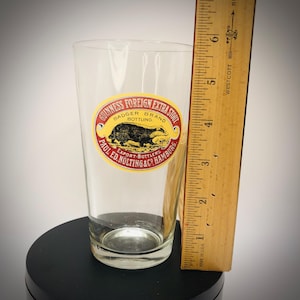K&ouml;nnte beinhalten: Ein klares Bierglas mit einem Guinness Foreign Extra Stout-Etikett mit einer Dachs-Illustration. Das Etikett ist gelb, rot und schwarz. Ein Holzlineal misst etwa 15 cm.