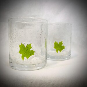 Puede incluir: Dos vasos de vidrio transparente con superficie texturizada. Cada vaso presenta un diseño de hoja de arce verde brillante. Los vasos son cilíndricos y parecen hechos a mano, con pequeñas burbujas.