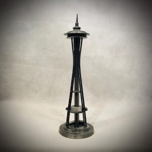 May include: A detailed metal replica of the Seattle Space Needle. The model features a dark, metallic finish with intricate structural details, including the observation deck and spire. The base is circular, and the overall design is a faithful representation of the iconic landmark.