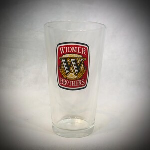 Puede incluir: Una taza de cerámica blanca con un asa negra y el texto "But First, Coffee" en negrita negra. La taza se encuentra sobre un fondo borroso, sugiriendo un ambiente acogedor.