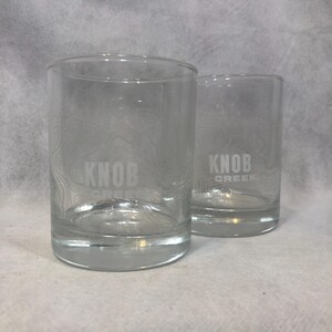 May include: Two clear glass tumblers featuring the etched text KNOB CREEK on the front. Each glass has a subtle, etched topographic map pattern surrounding the brand name, with a thick, solid glass base and straight, cylindrical sides.