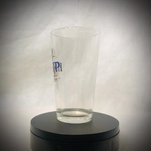 Puede incluir: Un vaso de pinta transparente con forma cil&iacute;ndrica. El vaso tiene un logotipo azul y blanco que dice "RPI" y "BERU". El vaso est&aacute; sobre un soporte giratorio negro.