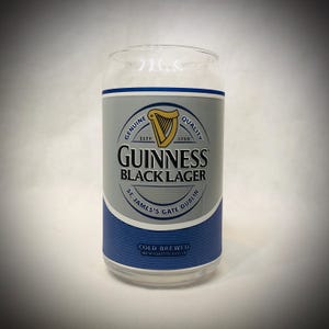 Może przedstawiać: Przezroczysty szklany kubek w kształcie puszki z logo Guinness Black Lager. Logo zawiera złotą harfę, napisy "Guinness Black Lager" i "St. James's Gate Dublin". Szklanka ma niebiesko-srebrną etykietę.