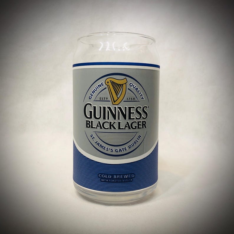 Może przedstawiać: Przezroczysty szklany kubek w kształcie puszki z logo Guinness Black Lager. Logo zawiera złotą harfę, napisy "Guinness Black Lager" i "St. James's Gate Dublin". Szklanka ma niebiesko-srebrną etykietę.