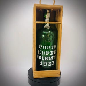 Puede incluir: Una botella de vidrio verde de Porto Kopke Colheita 1987 en una caja de madera. La botella tiene una etiqueta blanca y el texto "PORTO KOPKE COLHEITA 1987" en blanco. La caja de madera tiene un asa de cuerda.