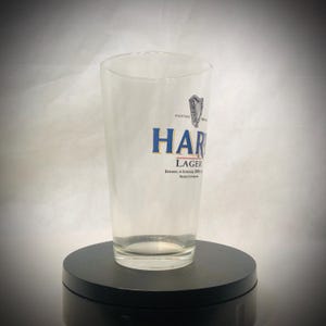 Puede incluir: Un vaso de pinta transparente con la palabra "HARP" en letras azules y doradas. El vaso tambi&eacute;n presenta la palabra "LAGER" y el texto "BREWED IN DUNDALK, IRELAND". Un logotipo de arpa es visible.