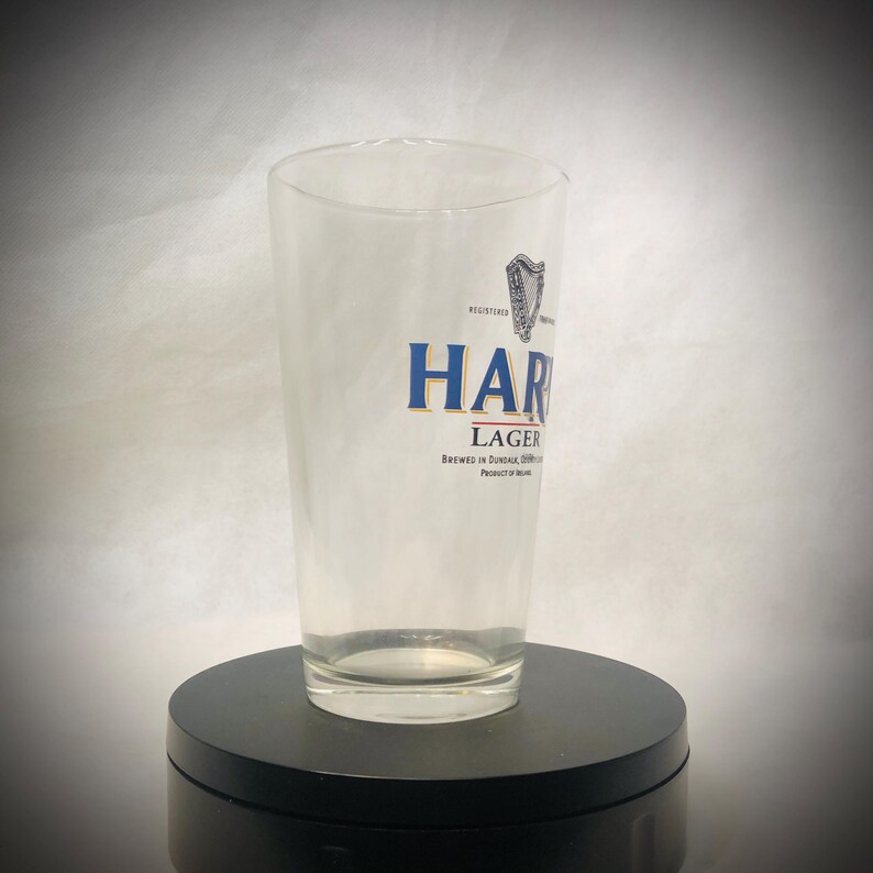 Puede incluir: Un vaso de pinta transparente con la palabra "HARP" en letras azules y doradas. El vaso tambi&eacute;n presenta la palabra "LAGER" y el texto "BREWED IN DUNDALK, IRELAND". Un logotipo de arpa es visible.