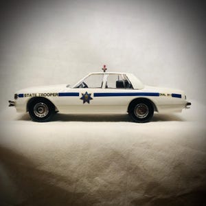 May include: A white toy car with blue stripes and the words "STATE TROOPER" and "DIAL 911" printed on the side. The car has a red light on top and a blue and gold emblem. The car is a vintage model.