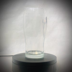 Puede incluir: Un vaso de pinta transparente y vac&iacute;o, con forma curvada, adecuado para cerveza. El vaso tiene un dise&ntilde;o sutil en el lateral y se apoya en una plataforma giratoria negra. El fondo es un degradado de blanco y gris.