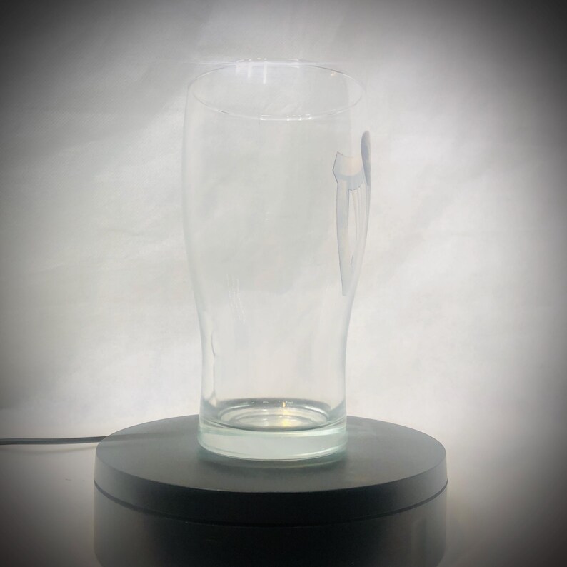 Puede incluir: Un vaso de pinta transparente y vac&iacute;o, con forma curvada, adecuado para cerveza. El vaso tiene un dise&ntilde;o sutil en el lateral y se apoya en una plataforma giratoria negra. El fondo es un degradado de blanco y gris.