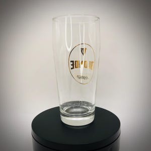 May include: A clear pint glass with a gold logo featuring the word "BROIDE" in a circular design. The glass is empty and sits on a black surface. The glass is designed for beverages.