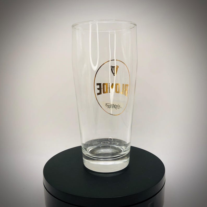 May include: A clear pint glass with a gold logo featuring the word "BROIDE" in a circular design. The glass is empty and sits on a black surface. The glass is designed for beverages.