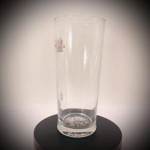 May include: A clear, tall glass with a slightly flared top. The glass has a logo in gold and a snowflake design on the bottom. The glass is empty and sits on a black surface.