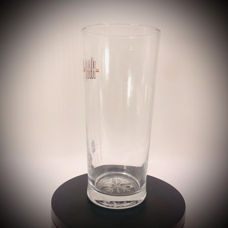 May include: A clear, tall glass with a slightly flared top. The glass has a logo in gold and a snowflake design on the bottom. The glass is empty and sits on a black surface.