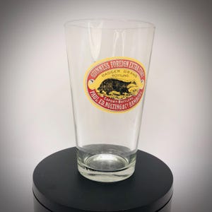K&ouml;nnte beinhalten: Ein klares Pintglas mit einem Guinness Foreign Extra Stout Badger Brand Etikett. Das ovale Etikett hat rote und gelbe Akzente und zeigt eine Dachs-Illustration. Das Glas ist leer und steht auf einer schwarzen Oberfl&auml;che.