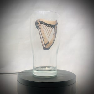Puede incluir: Un vaso de pinta transparente con un logotipo de arpa marr&oacute;n. El vaso es cil&iacute;ndrico con una parte superior ligeramente ensanchada. El logotipo del arpa est&aacute; centrado y tiene un aspecto vintage. El vaso est&aacute; sobre una base giratoria negra.