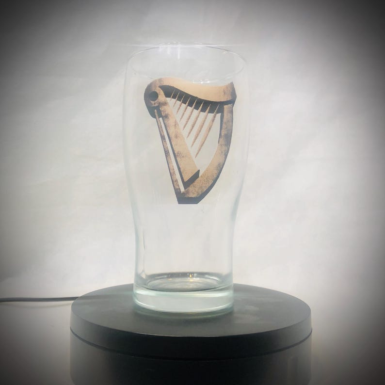 Puede incluir: Un vaso de pinta transparente con un logotipo de arpa marr&oacute;n. El vaso es cil&iacute;ndrico con una parte superior ligeramente ensanchada. El logotipo del arpa est&aacute; centrado y tiene un aspecto vintage. El vaso est&aacute; sobre una base giratoria negra.