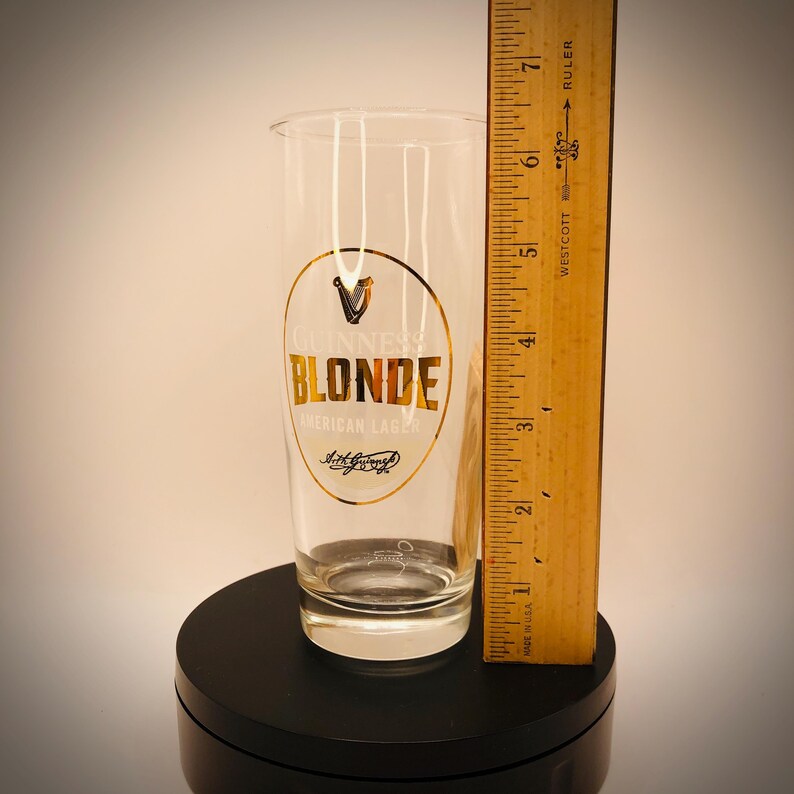 May include: A clear Guinness Blonde American Lager pint glass with gold lettering and a gold logo. The glass is approximately 7 inches tall and is on a black surface.