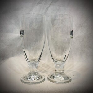May include: Two clear glass beer glasses with a stemmed base and a flared rim. Each glass features a partial black and red logo printed on the side. The glasses have a smooth, transparent surface and a sturdy circular foot for stability.