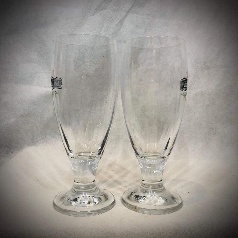 May include: Two clear glass beer glasses with a stemmed base and a flared rim. Each glass features a partial black and red logo printed on the side. The glasses have a smooth, transparent surface and a sturdy circular foot for stability.