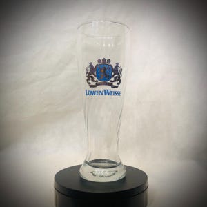 Puede incluir: Un vaso de cerveza alto y transparente con un diseño curvo. El vaso presenta un emblema azul y dorado con las palabras "Löwen Weisse" en letras azules. El vaso está diseñado para contener cerveza.