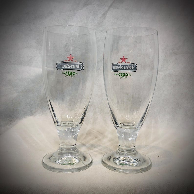 May include: Two clear glass beer glasses with a stemmed base. Each glass features the red star logo and the word Heineken printed in black text, accompanied by a green leaf graphic design centered on the front of the curved glass body.