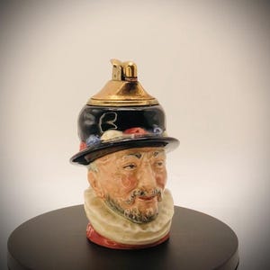 May include: A ceramic figurine of a man's head wearing a black hat with a gold top. The man has a white ruffled collar and a painted face with a beard. The hat has red, white, and blue accents.