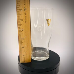 May include: A clear pint glass with the word "GUINNESS" etched on the front. A gold harp logo is visible. A wooden ruler is placed next to the glass. The glass measures approximately 5 inches in height.