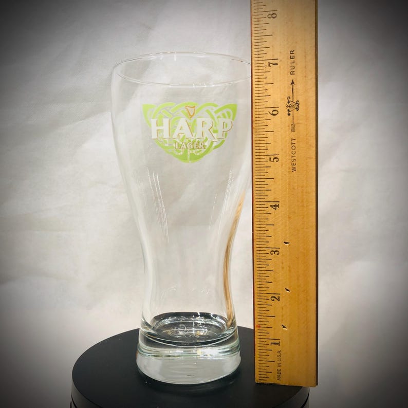 May include: A clear Harp Lager beer glass with a green and white logo. The glass is approximately 7 inches tall. A wooden ruler is placed next to the glass for scale. The glass is on a black surface.