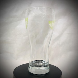 May include: A clear, tulip-shaped beer glass with a wide base and a narrow top. The glass has a green logo on both sides. The glass is empty and sits on a black surface.