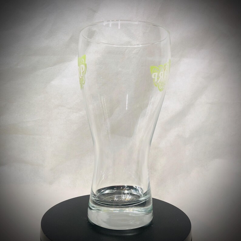 May include: A clear, tulip-shaped beer glass with a wide base and a narrow top. The glass has a green logo on both sides. The glass is empty and sits on a black surface.