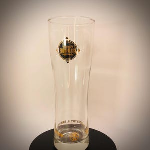 K&ouml;nnte beinhalten: Ein klares, hohes Bierglas mit leicht ausgestellter Form. Das Glas hat ein gold-schwarzes Logo mit dem Text "Blonde" und "Brewery & Brews". Das Glas ist zum Servieren von Bier bestimmt und hat einen wei&szlig;en Sockel.