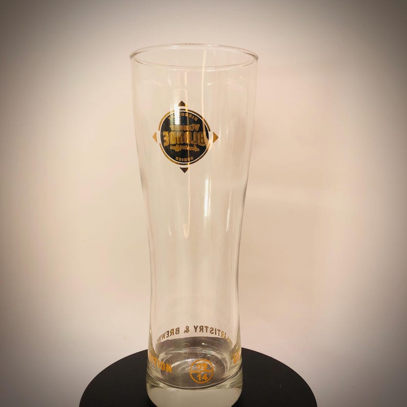 K&ouml;nnte beinhalten: Ein klares, hohes Bierglas mit leicht ausgestellter Form. Das Glas hat ein gold-schwarzes Logo mit dem Text "Blonde" und "Brewery & Brews". Das Glas ist zum Servieren von Bier bestimmt und hat einen wei&szlig;en Sockel.