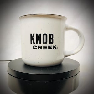 Puede incluir: Taza de cerámica blanca con diseño moteado negro y borde negro. La taza presenta el texto "KNOB CREEK" en letras negras en negrita. La taza tiene un asa curva y probablemente se usa para bebidas calientes.