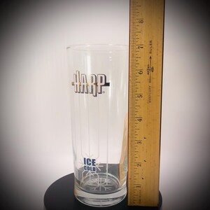 May include: A clear glass with the word "HARP" in gold and black lettering. The glass has vertical lines and the words "ICE COLD" in blue. A wooden ruler is next to the glass, measuring approximately 7 inches.