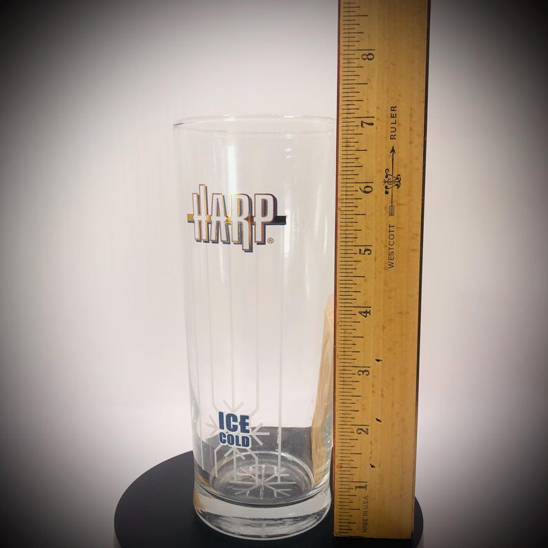 May include: A clear glass with the word "HARP" in gold and black lettering. The glass has vertical lines and the words "ICE COLD" in blue. A wooden ruler is next to the glass, measuring approximately 7 inches.