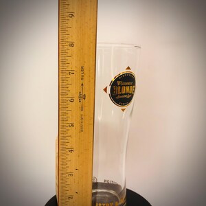 K&ouml;nnte beinhalten: Ein klares Guinness Blonde American Lager Bierglas mit einem gold-schwarzen Logo. Das Glas ist etwa 18 cm hoch, gemessen mit einem Holzlineal. Das Glas steht auf einer schwarzen Oberfl&auml;che.